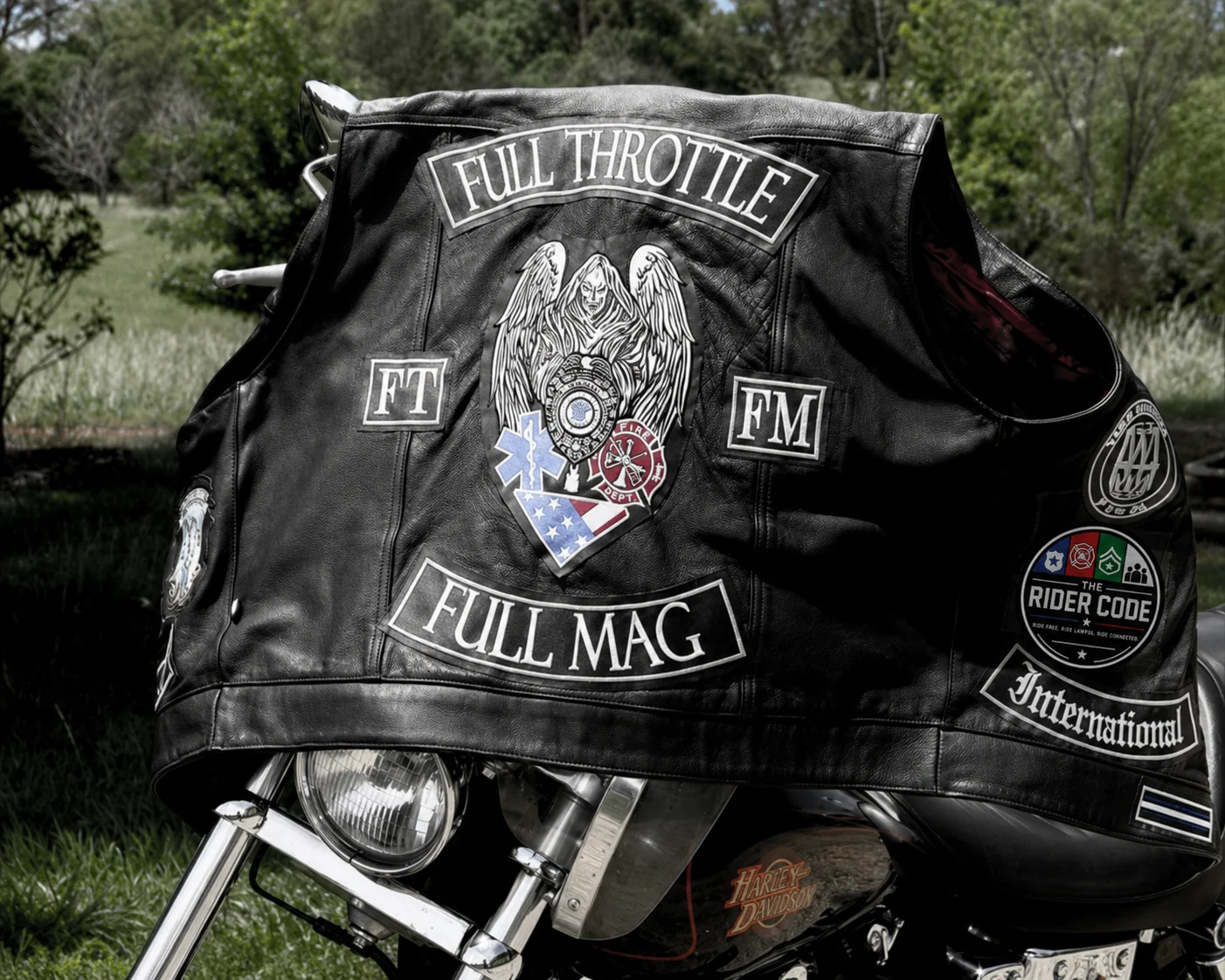 An FTFM leather vest draped over a Harley, showing FULL THROTTLE and FULL MAG rockers, the FT/FM patches, the International rocker, and The Rider Code emblem.